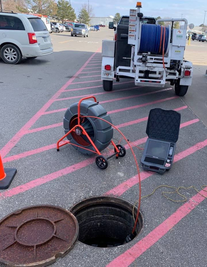 Picture of a hydro-jetter trailer in front of an open manhole with a pipe televising camera rig leading to a long chord going into the manhole