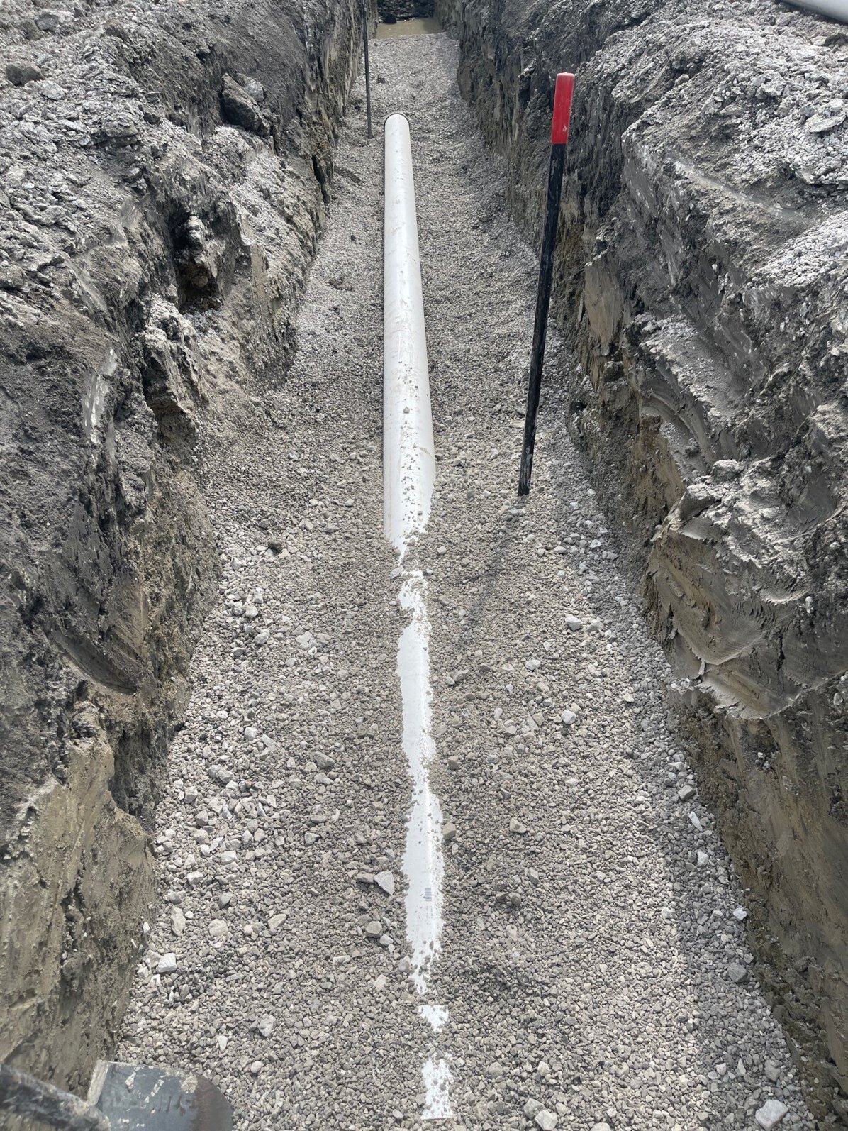 A large commercial sewer installation: The picture shows a rectangular excavation with a new, large white PVC pipe set in gravel with a red capped metal meauring stake that shows the excavator proper depth.