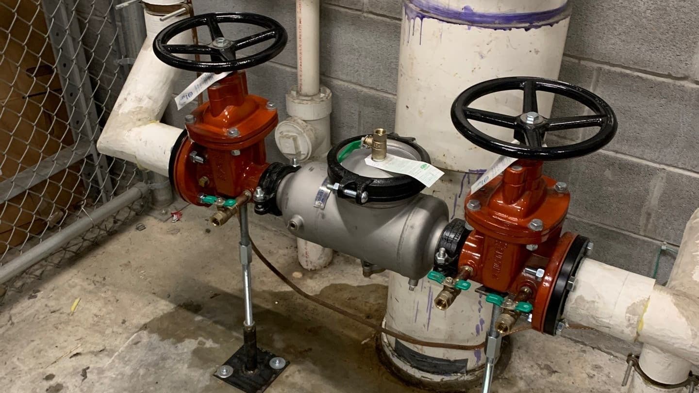 Picture shows a newly installed back-flows prevention assembly on a large industrial water main inside a facility. There is grey cinderblock wall behind, concrete floor beneath, and a fenced off area on the right side of the phone. The piping is descending form the ceiling, turns 90 degrees to run parallel to the ground for 4 feet, around 2 feet high, and then ascends to the ceiling again, and a new back-flow prevention assembly has been installed in that parallel part of the piping.