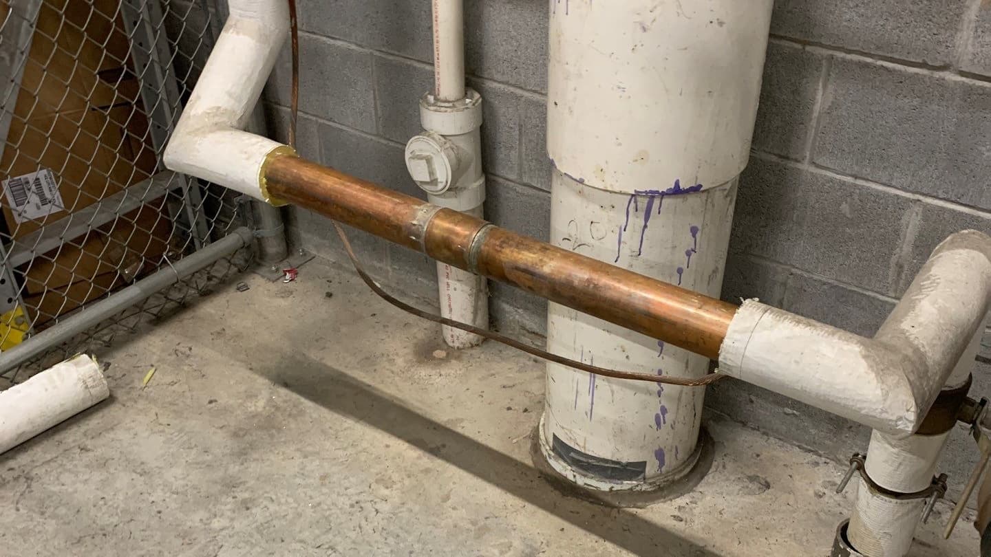 Picture shows a large industrial water main inside a facility. There is grey cinderblock wall behind, concrete floor beneath, and a fenced off area on the right side of the phone. The piping is descending form the ceiling, turns 90 degrees to run parallel to the ground for 4 feet, around 2 feet high, and then ascends to the ceiling again.