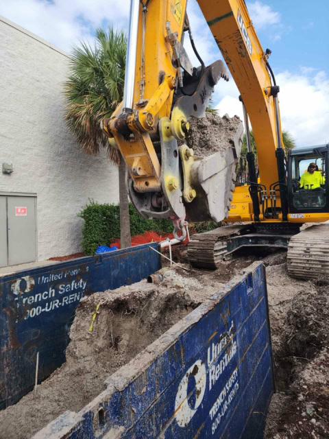 An excavator operator is using an excavator to dig within a trench box. There is an large commercial building in the background.